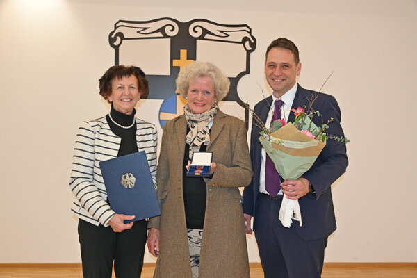 Die stellvertretende Landrätin Notburga Kunert, Christine von Gadow und Bürgermeister Sven Schnieber stehen mit Urkunde, Medaille und Blumenstrauß in den Händen vor dem Stadtwappen.