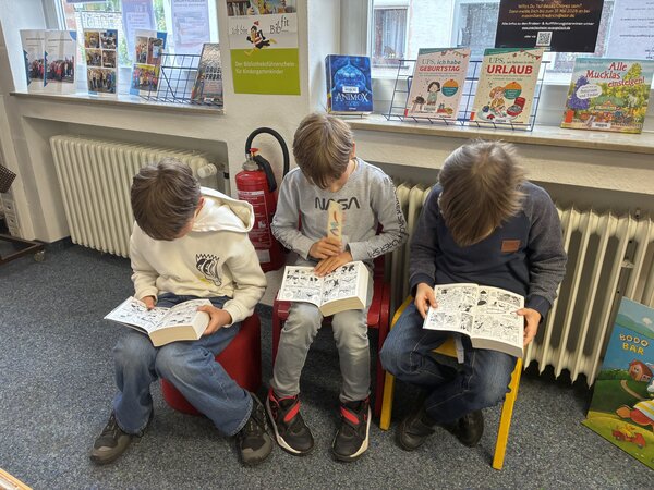 Drei Kinder sitzen auf zwei Stühlen und einem Hocker und haben auf ihren Beinen Mangas aufgeschlagen.