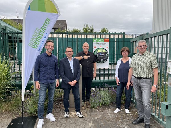 Die Beteiligten stehen am Werkstor, an dem der neue Defibrillator angebracht ist.
