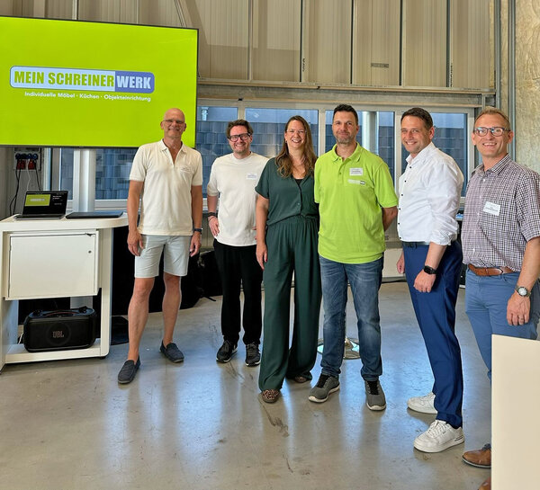 Die Protagonistinnen und Protagonisten der Veranstaltung stehen vor einem Monitor mit Schriftzug des Unternehmens.
