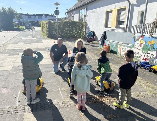 Die Verkehrssicherheitstrainer und Kinder befinden sich mit den Bobbycars auf einem kleinen Platz abseits der Straße.
